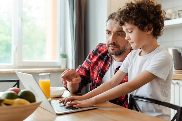 Foco seletivo de belo pai freelancer apontando com o dedo para laptop perto de filho encaracolado e café da manhã na mesa — Fotografia de Stock