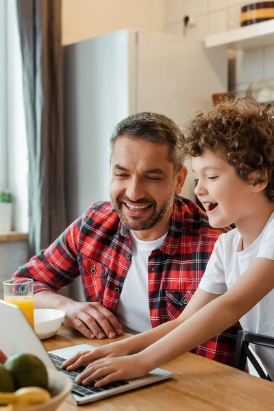 Enfoque selectivo de hijo rizado utilizando el ordenador portátil cerca de padre freelancer feliz y desayuno en la mesa - foto de stock