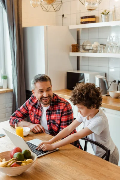 Curly boy using laptop near happy freelancer father and breakfast on table — Stock Photo