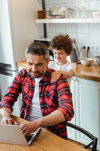 Selective focus of handsome freelancer father using laptop near curly son — Stock Photo