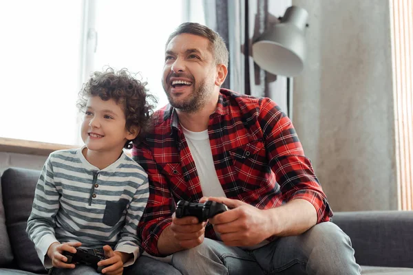 KIEW, UKRAINE - 14. MAI 2020: fröhlicher Vater und lockiger Sohn beim Videospiel im Wohnzimmer — Stockfoto