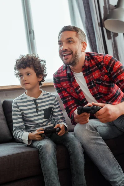 KIEW, UKRAINE - 14. MAI 2020: Glücklicher Vater und lockiger Sohn beim Videospiel im Wohnzimmer — Stockfoto