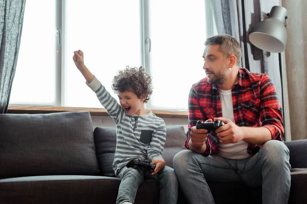 KYIV, UKRAINE - 14 MAI 2020 : beau père regardant le fils bouclé excité célébrer le triomphe tout en jouant au jeu vidéo dans le salon — Photo de stock