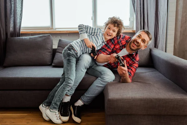 KYIV, UCRANIA - 14 de mayo de 2020: padre feliz e hijo alegre jugando videojuegos en la sala de estar - foto de stock