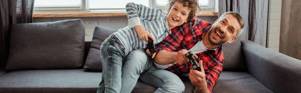 KYIV, UCRANIA - 14 de mayo de 2020: concepto panorámico de padre guapo e hijo feliz jugando videojuegos en la sala de estar - foto de stock