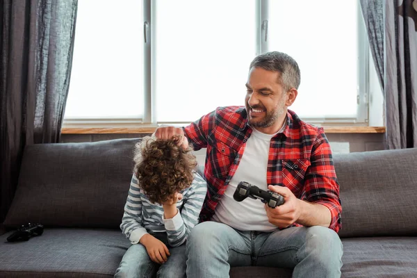 KYIV, UKRAINE - MAY 14, 2020: happy father touching upset and curly son  while playing video game in living room — Stock Photo