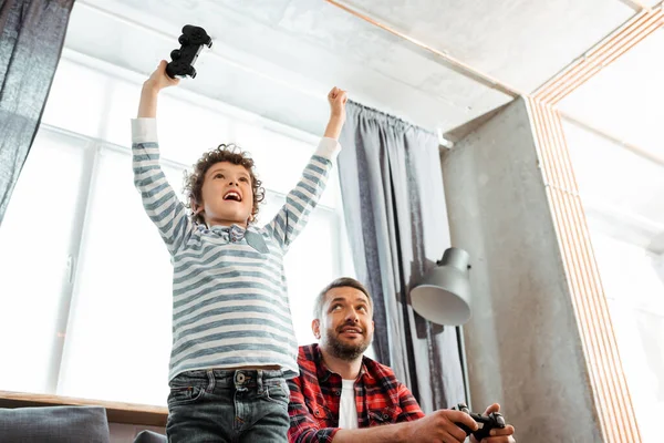 KYIV, UKRAINE - MAY 14, 2020: low angle view of handsome father looking at excited curly son celebrating triumph while playing video game — Stock Photo
