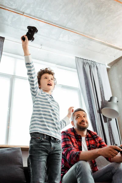 KYIV, UCRANIA - 14 de mayo de 2020: vista de bajo ángulo del padre guapo mirando al hijo emocionado celebrando el triunfo mientras juega a videojuegos - foto de stock