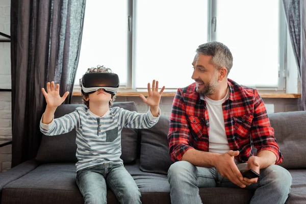 Счастливый отец держит пульт дистанционного управления и смотрит на кудрявого сына в гарнитуре виртуальной реальности — стоковое фото
