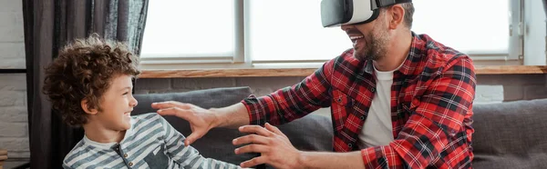 Cosecha horizontal de padre alegre en auriculares de realidad virtual gesto cerca de hijo feliz - foto de stock