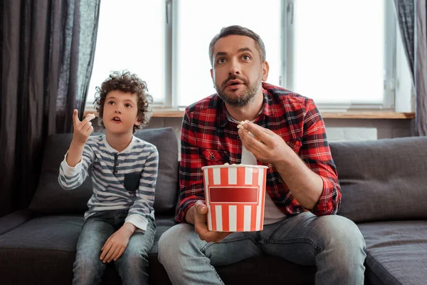 Foco seletivo do pai segurando balde de pipoca perto de filho encaracolado apontando com o dedo enquanto assiste filme — Fotografia de Stock