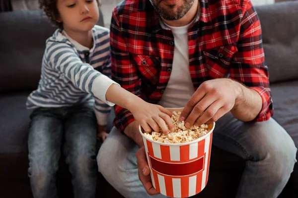 Vista ritagliata del bambino raggiungendo gustosi popcorn vicino padre barbuto — Foto stock
