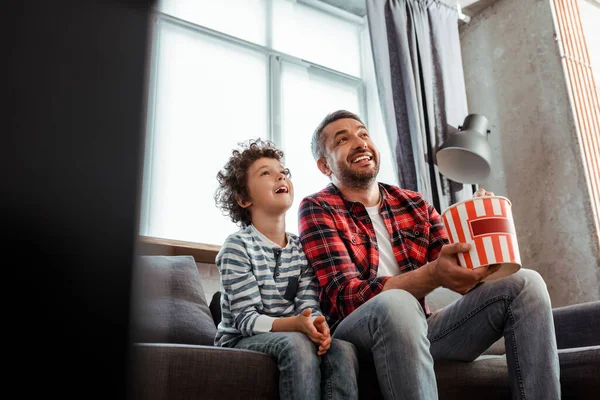 Selektiver Fokus des fröhlichen und lockigen Kindes, das Film in der Nähe des Vaters mit Popcorn-Eimer anschaut — Stockfoto