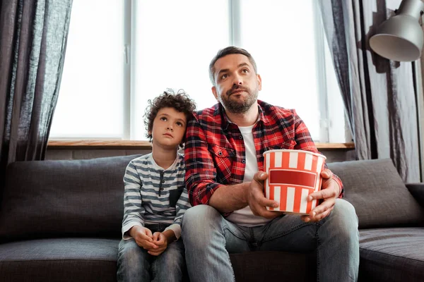 Кудрявый ребенок смотрит фильм рядом с бородатым отцом, держащим ведро попкорна — стоковое фото