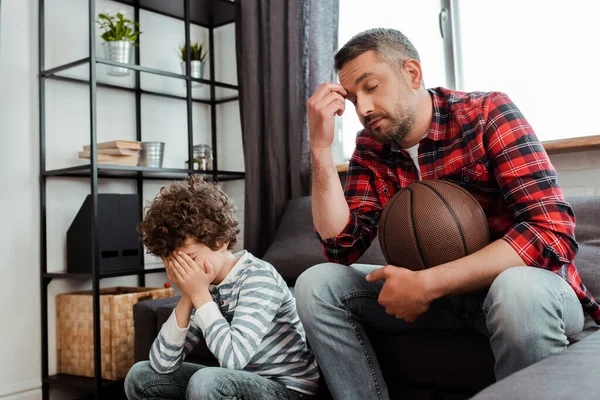 Displeased man holding basketball while watching championship with upset son covering face — Stock Photo