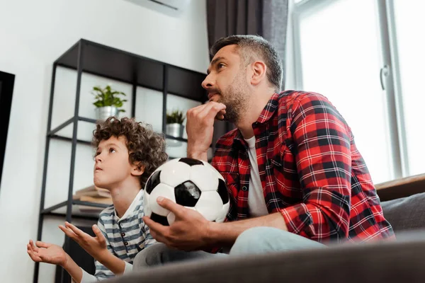 Selective focus of bearded father holding football and watching championship with curly son — Stock Photo