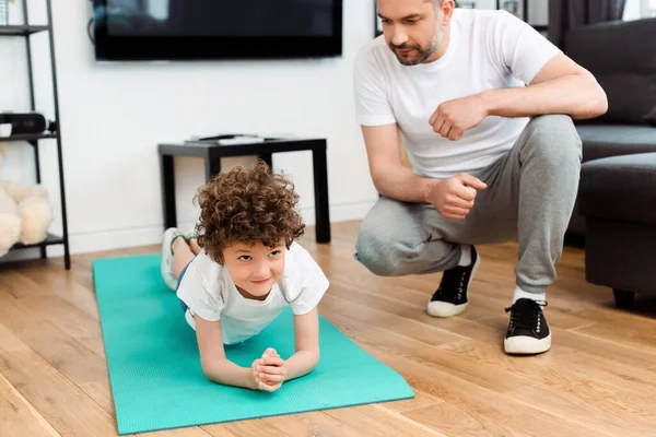 Père barbu regardant fils bouclé exercice sur tapis de fitness — Photo de stock