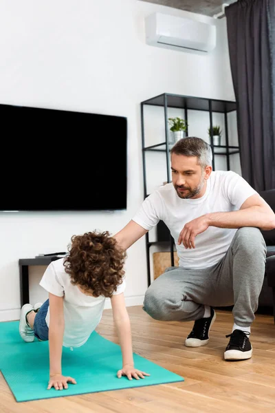 Bello padre guardando riccio figlio lavorare fuori su fitness mat — Foto stock