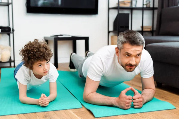 Bonito pai e encaracolado filho fazendo prancha em tapetes de fitness — Fotografia de Stock