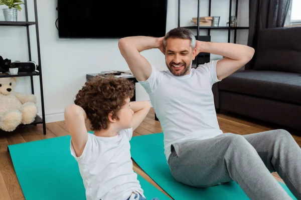 Lockiges Kind und bärtiger Vater schauen einander beim Training auf Fitnessmatten zu Hause an — Stockfoto