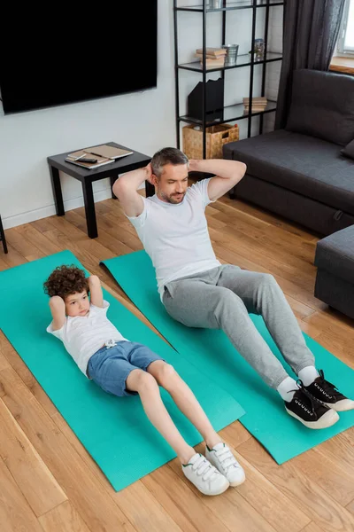 Niño rizado y padre barbudo haciendo ejercicio en colchonetas de fitness en casa - foto de stock