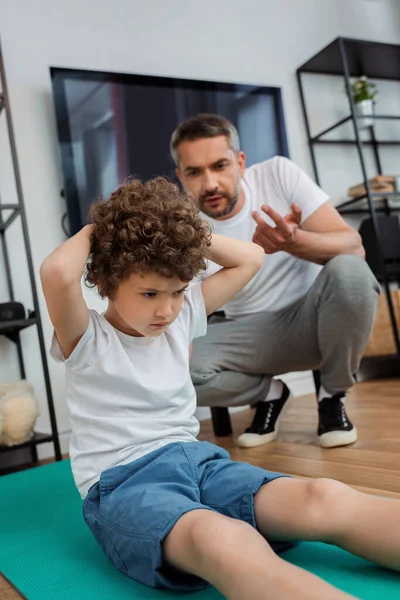 Messa a fuoco selettiva del bambino riccio che lavora sul tappeto fitness vicino al padre contando a casa — Foto stock