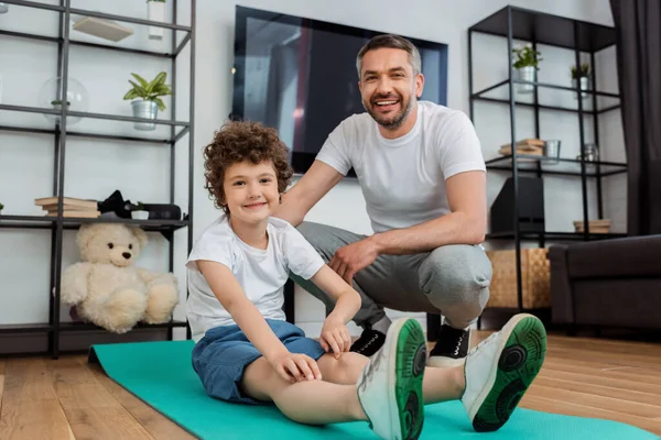 Fröhlicher Mann lächelt neben glücklichem Sohn auf Fitnessmatte — Stockfoto