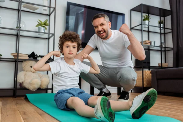 Glücklicher Vater jubelt neben lockigem Sohn und zeigt Muskeln — Stockfoto
