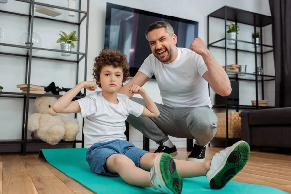 Feliz padre animando cerca rizado hijo mostrando músculo - foto de stock