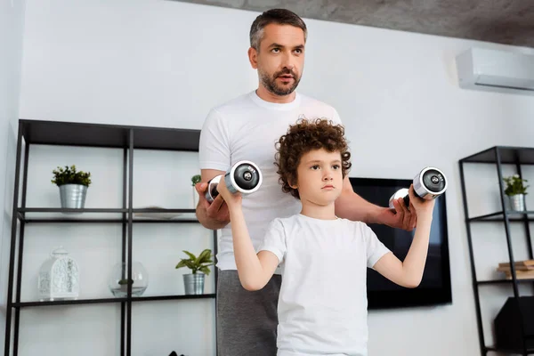 Padre guapo ayudando al hijo rizado a hacer ejercicio con pesas - foto de stock