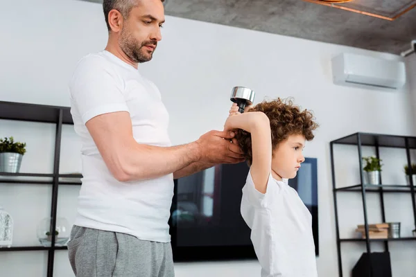 Бородатый отец трогает гантель, когда кудрявый сын тренируется дома — стоковое фото