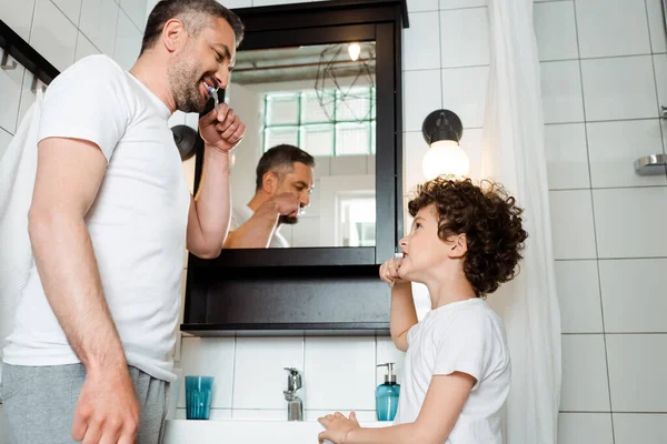 Padre guapo y rizado hijo cepillarse los dientes en el baño - foto de stock