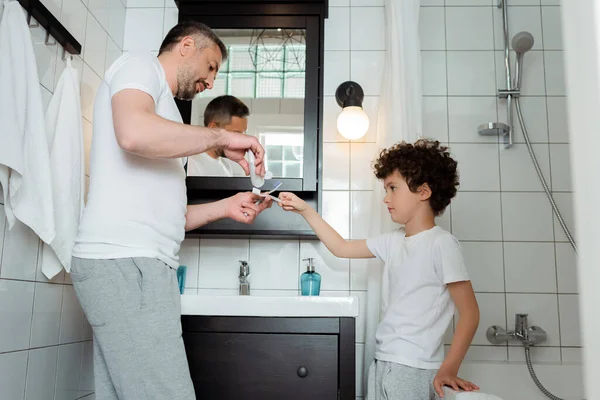 Schöner Vater hält Zahnpasta in der Nähe der Zahnbürste seines lockigen Sohnes — Stockfoto