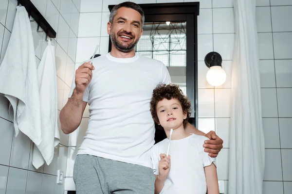 Alegre padre e hijo sosteniendo cepillos de dientes en el baño - foto de stock