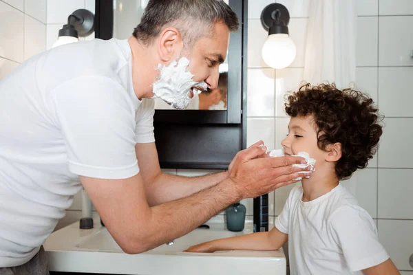 Vista lateral de feliz padre aplicando espuma de afeitar en la cara de lindo hijo en el baño - foto de stock