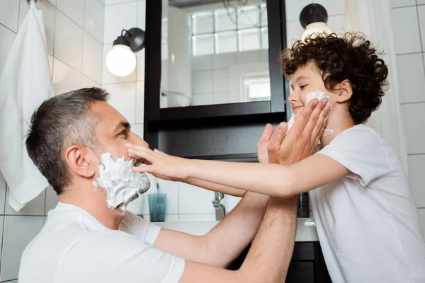 Vista lateral de feliz padre y lindo hijo con espuma de afeitar tocando caras en el baño - foto de stock