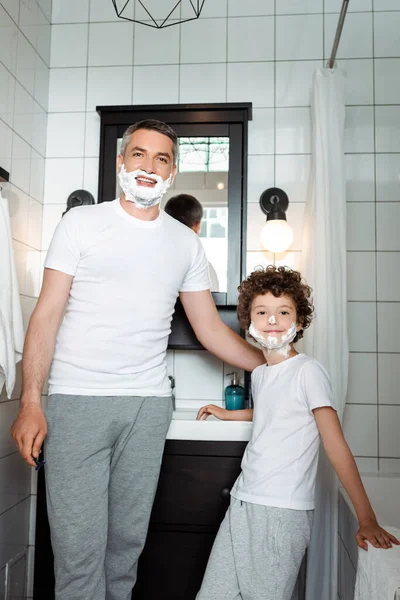 Happy father and curly son with shaving foam on faces looking at camera in bathroom — Stock Photo