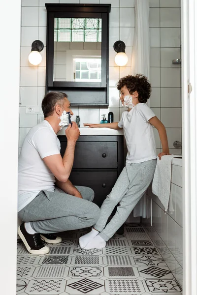 Schöner Vater mit Rasierschaum im Gesicht, der Rasiermesser hält, während er sich in der Nähe des lockigen Sohnes rasiert — Stockfoto