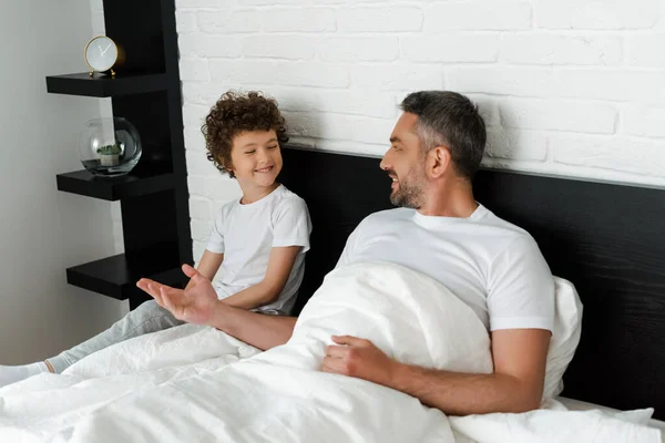Счастливый отец и кудрявый сын смотрят друг на друга и улыбаются в спальне — стоковое фото