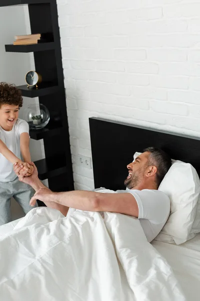 Niño rizado cogido de la mano mientras juega con el padre feliz en el dormitorio - foto de stock