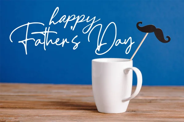 White cup and black decorative paper fake mustache on wooden surface isolated on blue, happy fathers day illustration — Stock Photo
