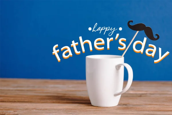 White cup and black decorative paper fake mustache on wooden surface isolated on blue, happy fathers day illustration — Stock Photo