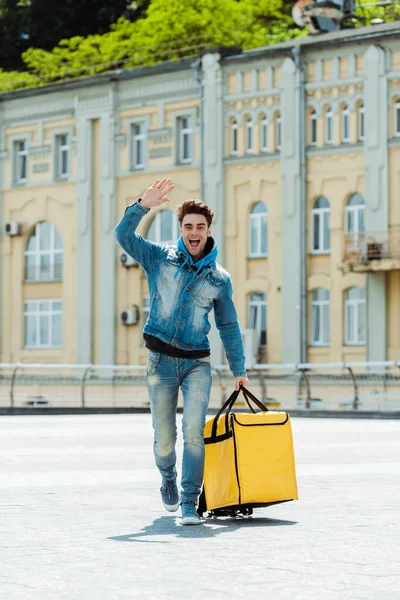 Mensajero positivo con bolsa térmica agitando la mano en la calle urbana - foto de stock