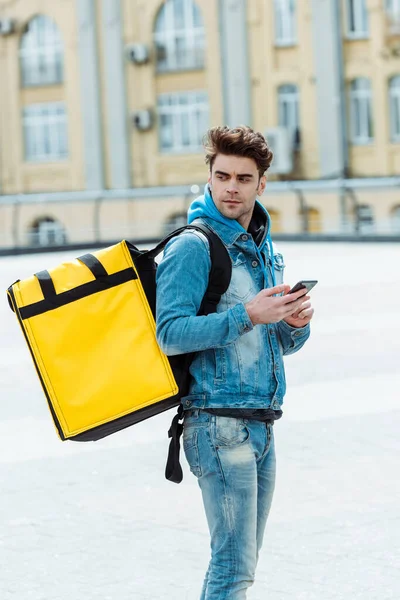 Side view of handsome courier with thermo backpack holding smartphone and looking away on urban street — Stock Photo