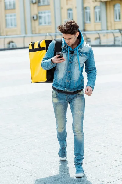 Handsome courier with thermo bag using cellphone while walking on urban street — Stock Photo