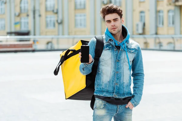 Handsome courier with thermo bag showing smartphone and looking at camera on city street — Stock Photo