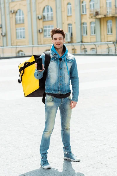 Hombre de reparto guapo con mochila termo mostrando teléfono inteligente y sonriendo a la cámara en la calle urbana - foto de stock