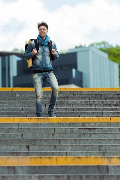 Selective focus of delivery man with thermo backpack smiling at camera on stairs on urban street — Stock Photo