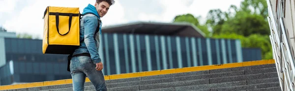 Panoramic crop of smiling courier with thermo backpack smiling at camera on stairs — Stock Photo