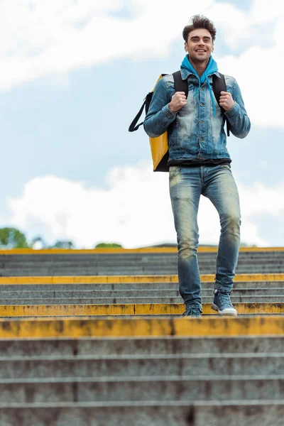 Low angle view of positive courier with thermo backpack walking on stairs with cloudy sky at background — Stock Photo